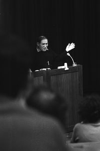 A person stands at a wooden podium, gesturing with one hand raised while speaking. The background is dark, and several audience members are visible in the foreground.
