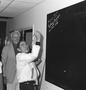 A man smiles while writing on a blackboard with chalk as another man stands beside him, also smiling. The setting appears to be indoors, possibly in an academic or professional environment.