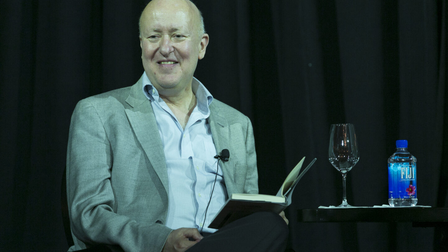 Man sitting on a stage as he holds a book on his leg