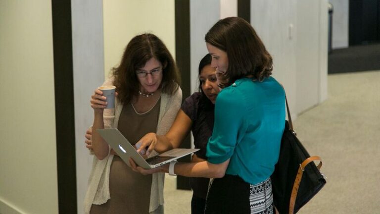 Three women standing and looking at a laptop that a woman on the right is holding