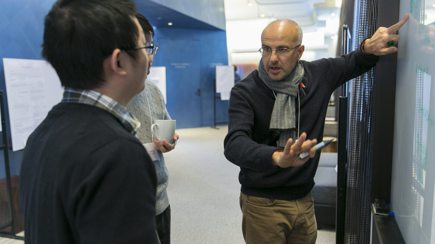Two people listening to a man pointing to green writing on a whiteboard