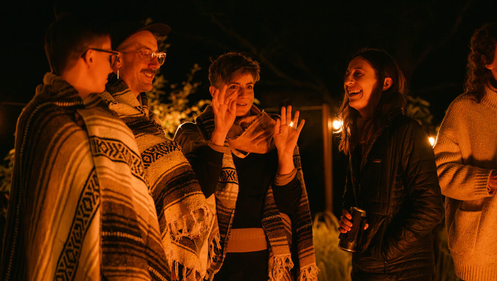 Four people laugh and talk around a campfire at night, wrapped in blankets and jackets, warmly lit by the fire; another person is partly visible on the right.