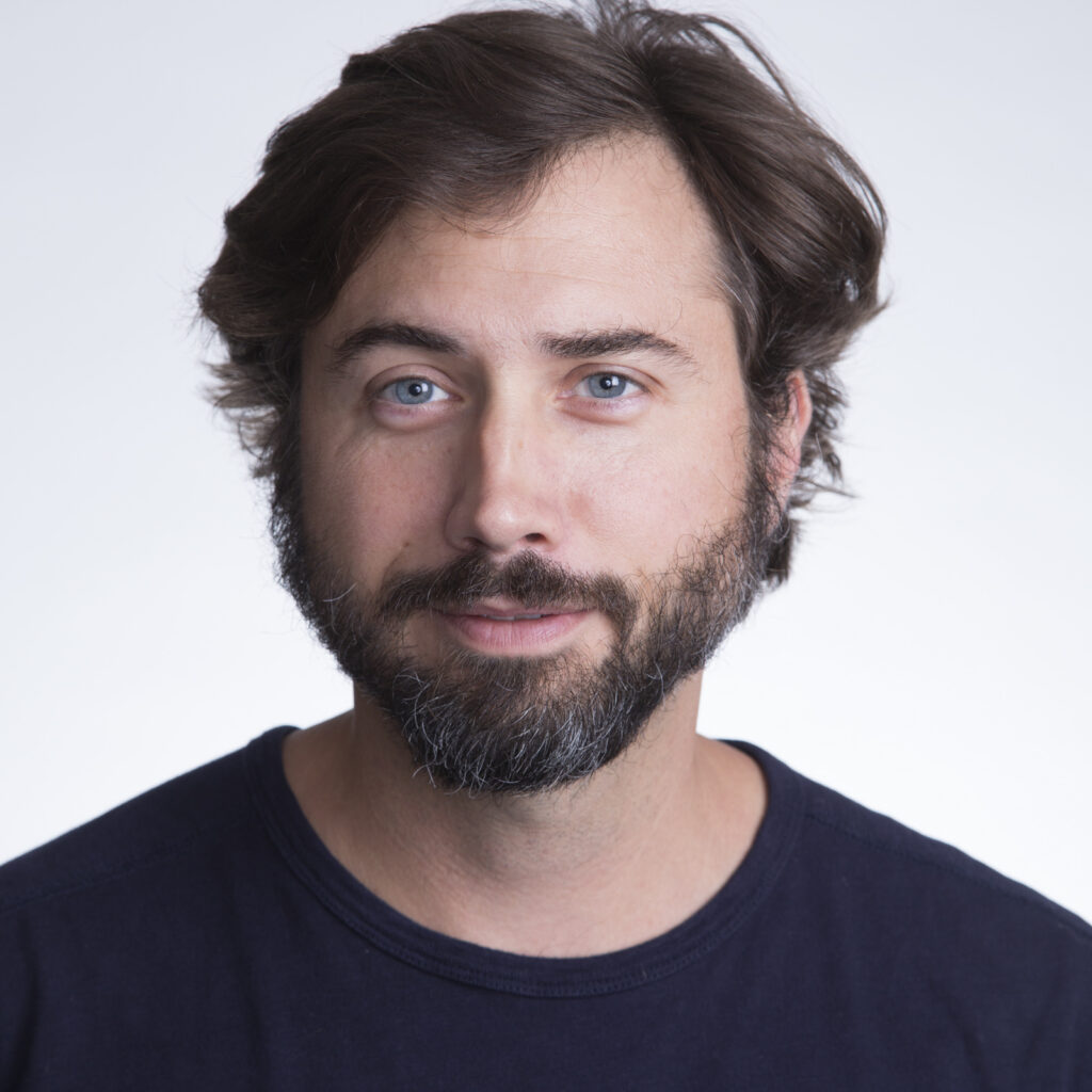 A man with light skin, brown hair, and a beard wearing a dark shirt stands against a plain light background, looking at the camera with a neutral expression.