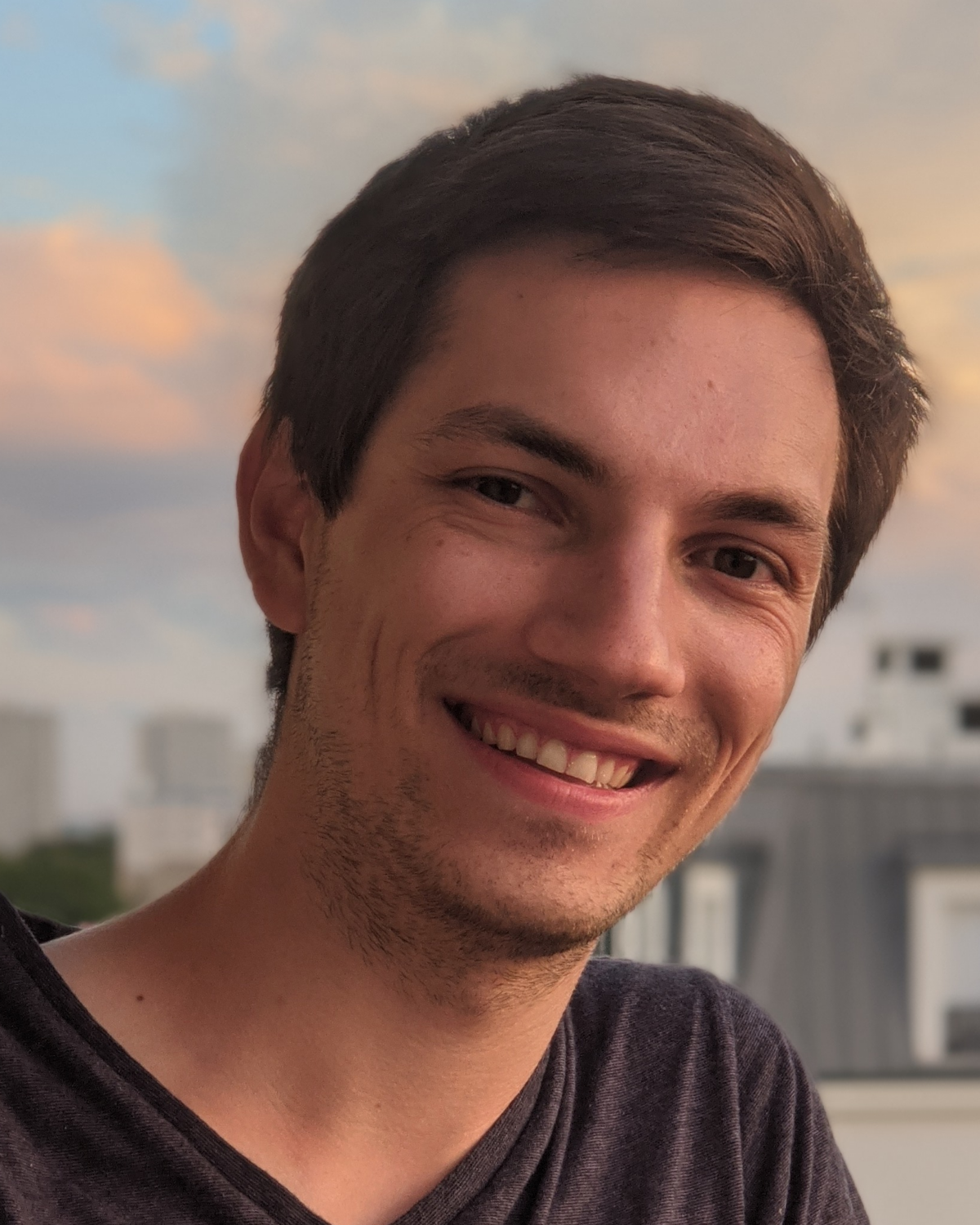 A young man with short brown hair smiles at the camera outdoors, with a blurred cityscape and sunset sky in the background.
