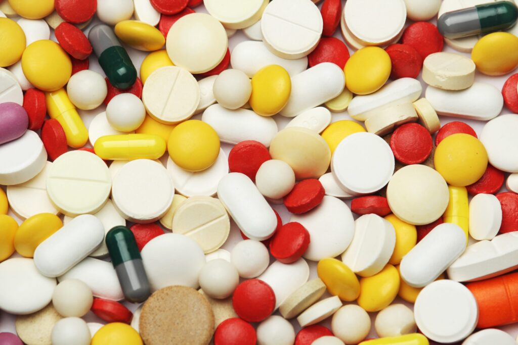 A large assortment of colorful pills and tablets, including white, yellow, red, green, and orange, are scattered in a close-up view.
