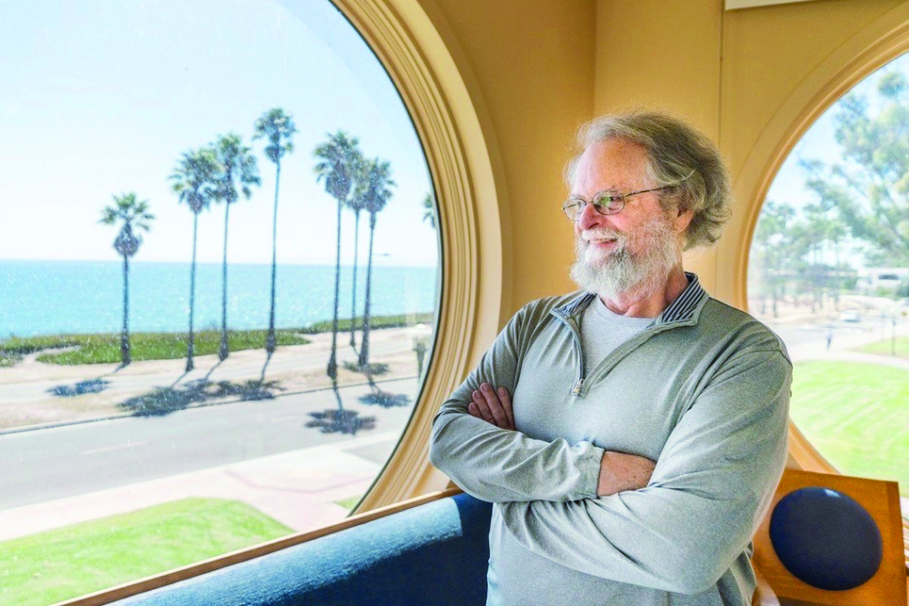 Bill Paxton stands in front of a window at the Kavli Institute for Theoretical Physics. Outside the window is the Pacific Ocean, a road and palm trees.