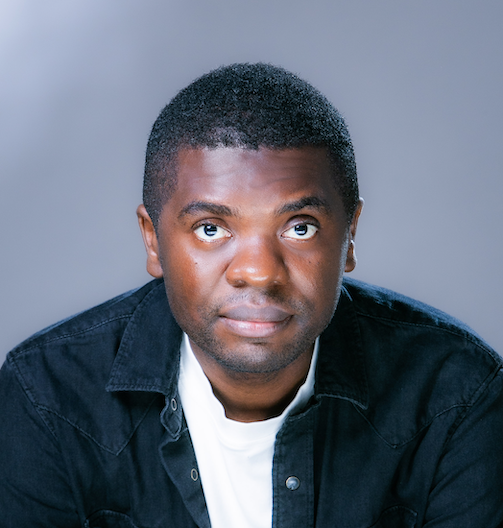 A man wearing a dark denim jacket over a white shirt looks upward slightly, set against a soft, blue-grey background.