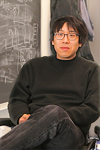 A person with glasses, wearing a dark sweater and jeans, sits with legs crossed in front of a chalkboard covered in diagrams and formulas.