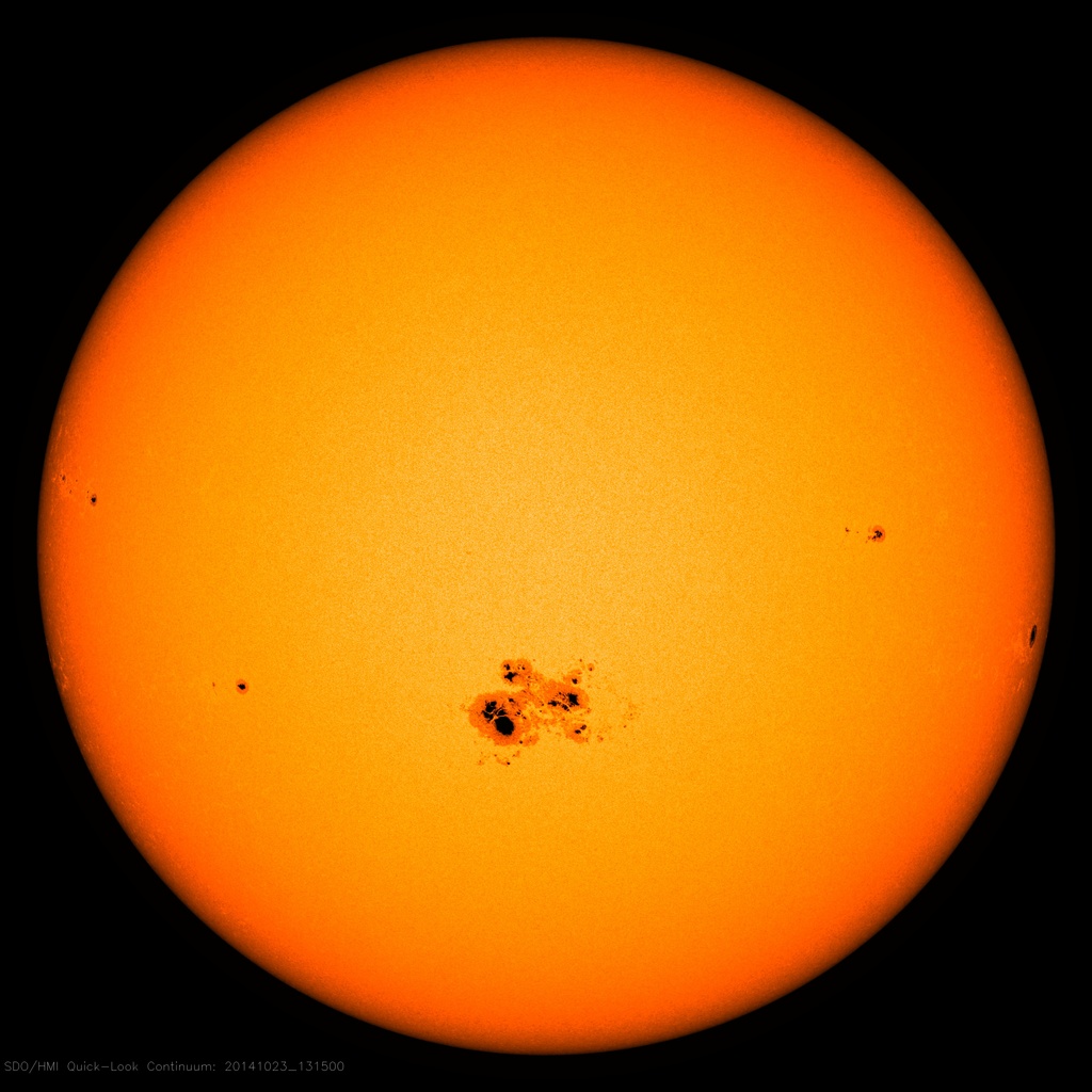 A bright orange image of the Sun shows several dark sunspots clustered near its center and scattered across its glowing surface, set against a black background.