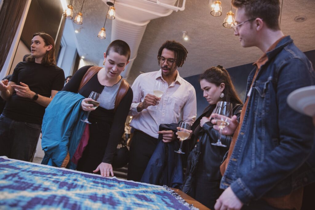 Five people stand around a table, holding drinks and looking at a blue-patterned textile. They appear engaged and interested, with warm lighting and modern decor in the background.