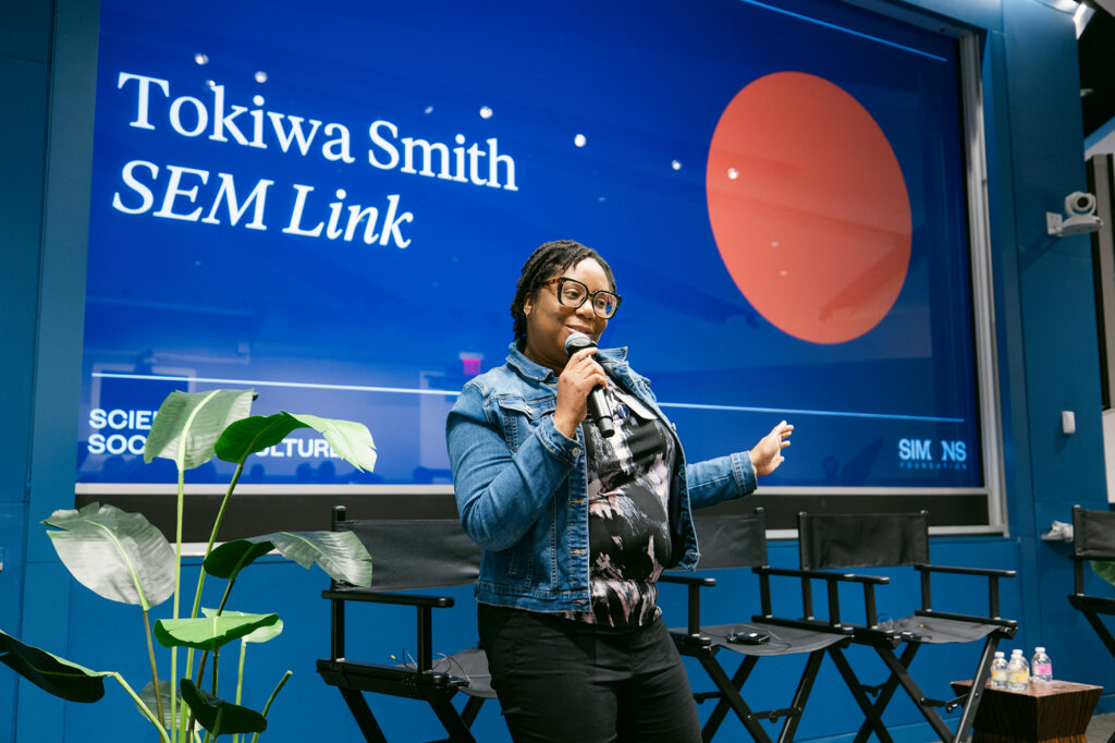 Black woman with glasses holding a microphone standing in an auditorium.