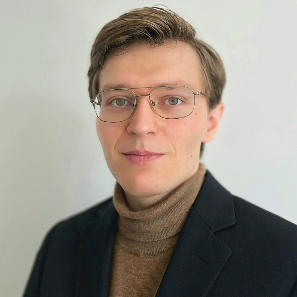 A person with short light brown hair, wearing glasses, a brown turtleneck sweater, and a dark blazer, stands against a plain light background, looking at the camera with a neutral expression.