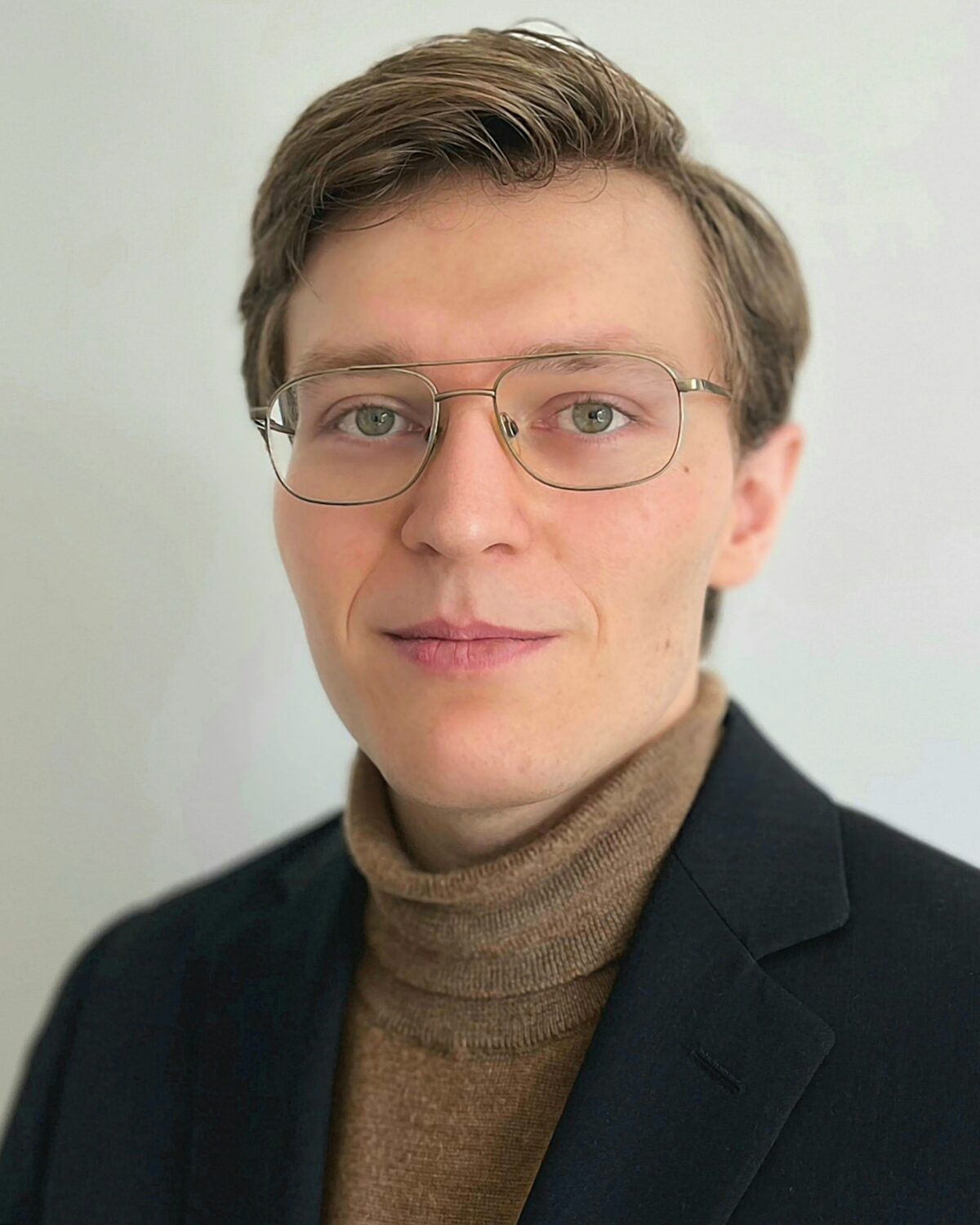 A person with short light brown hair, wearing glasses, a brown turtleneck sweater, and a dark blazer, stands against a plain light background, looking at the camera with a neutral expression.