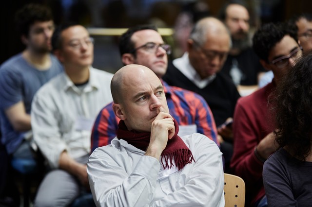 Man sitting in a lecture hall full of people as he puts his right hand up to his chin