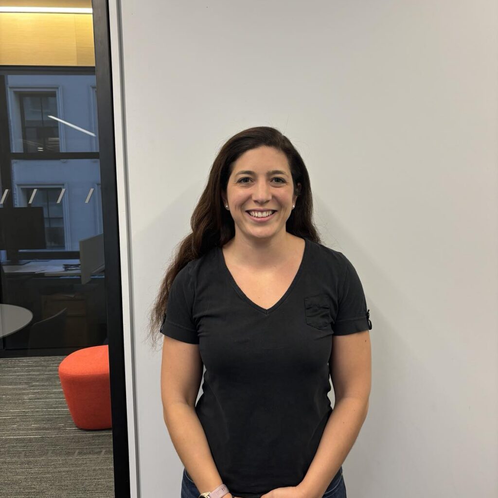 A woman with long brown hair wearing a black t-shirt and jeans stands smiling in front of a white wall, with an office space visible to the left.