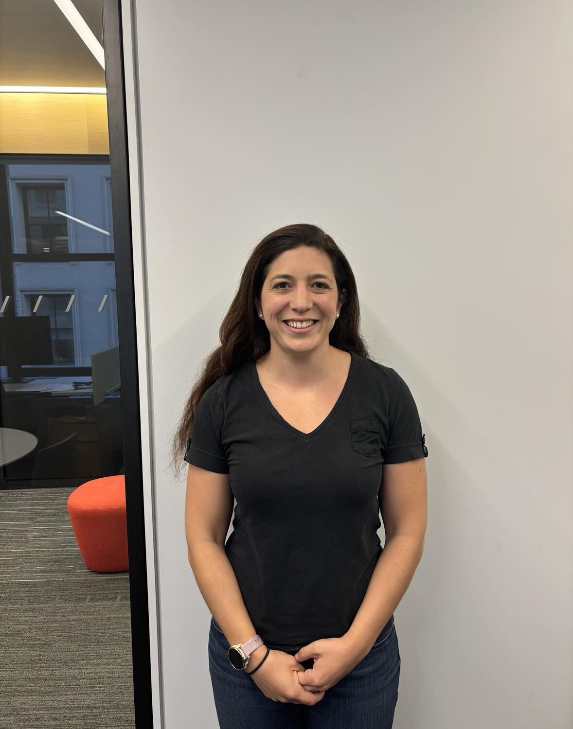 A woman with long brown hair wearing a black t-shirt and jeans stands smiling in front of a white wall, with an office space visible to the left.