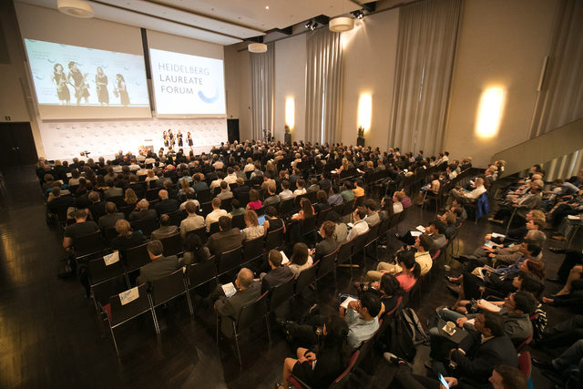 Large audience seated in a conference hall facing a stage with speakers. Two projection screens display the events logo and images. The room is well-lit, with tall curtains and people attentively watching the presentation.