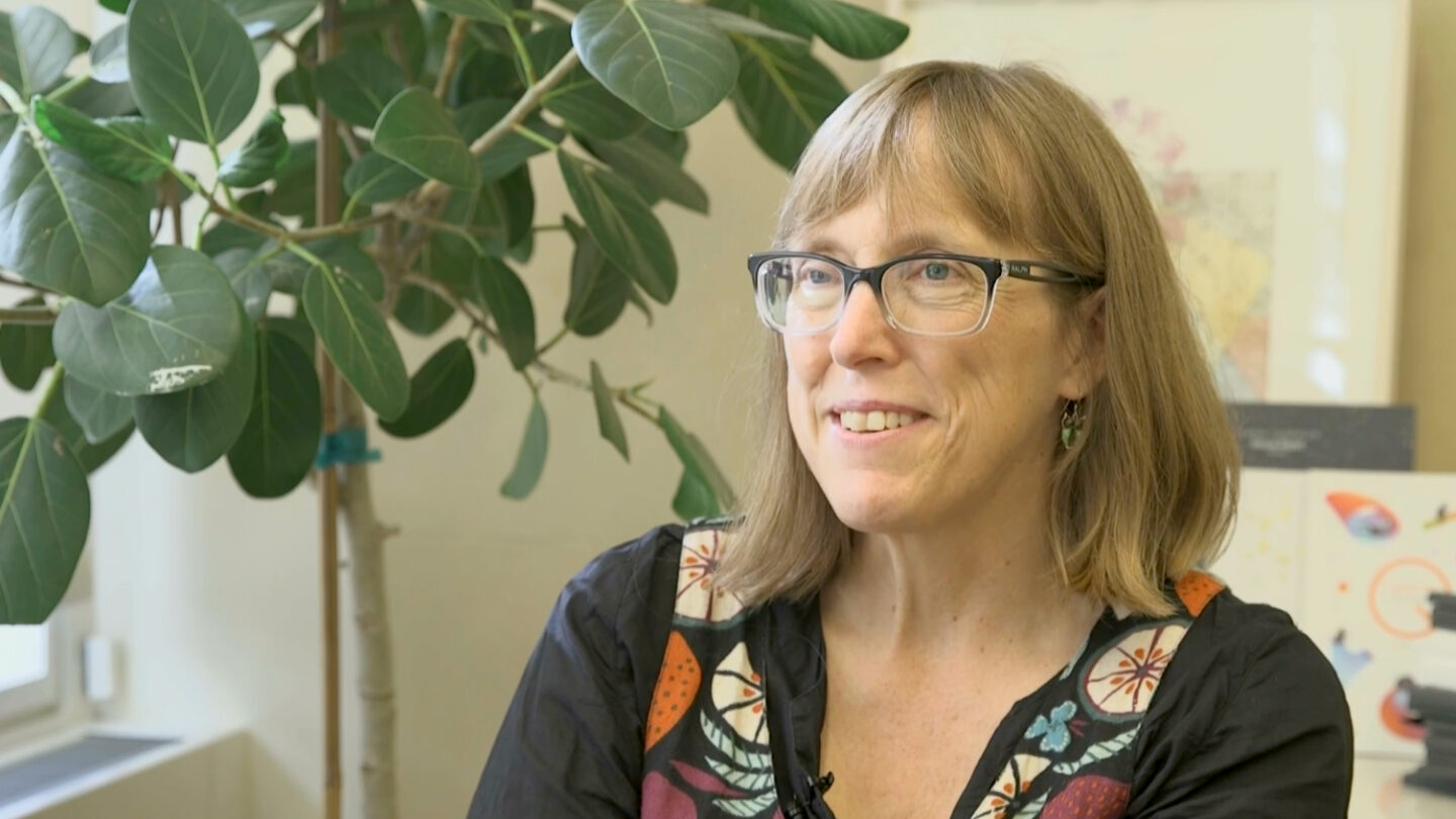A woman with glasses and shoulder-length blonde hair smiles while sitting indoors near a leafy plant. She is wearing a black top with colorful patterns.