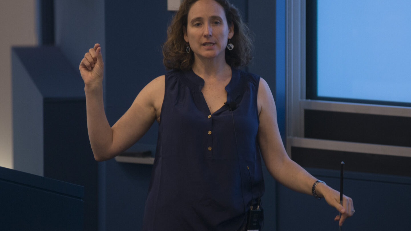 Woman standing by a lectern, as she gesticulates with her right hand