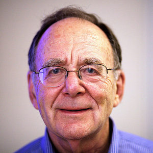 Older man with glasses, wearing a blue collared shirt, smiling gently at the camera with a neutral background.