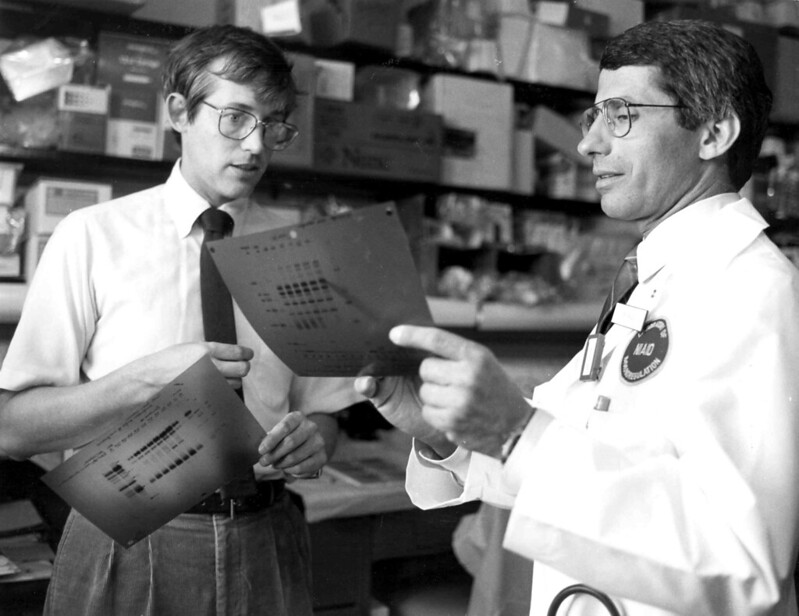 A black and white photo of a young Anthony Fauci and Clifford Lane. Fauci, on the left, has short combed-over dark hair, wireframe glasses, a white button-up shirt, black tie, and slacks with a belt. He is holding a three-holed chart in his left hand as he holds his tie with his left and looks towards Lane. Lane, on the right, has dark hair, wireframe glasses, a dark shirt, a white scientist's coat, a circular badge that visibly reads 