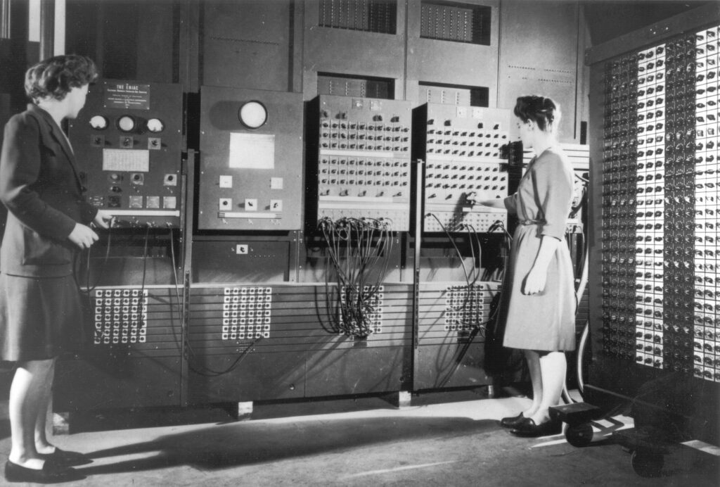A black-and-white photo of two of the original programmers for the ENIAC, with Jean Bartik being on the left and Frances Spence on the right, standing in front of an early model of a electronic computer, which resembles a screenless wooden cabinet with switches, buttons, ports, and wires.