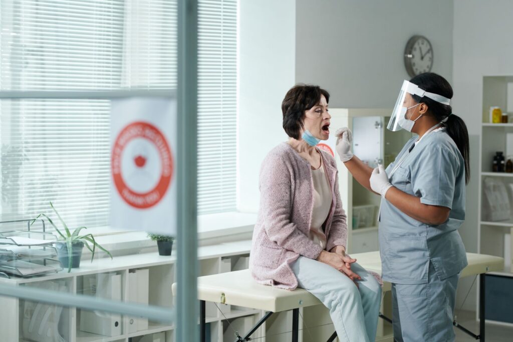A photograph of a nurse treating a female patient at a doctor's office. The female patient is sitting on a table, has a prodecure mask pulled down past her chin, and has her mouth agape. The nurse is wearing both a procedure mask and a face-shield as she appears to be putting a cotton swab into the patient's open mouth.
