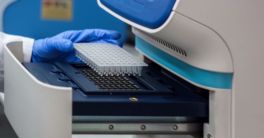 A photograph of DNA samples being placed in a PCR Machine by a gloved hand.