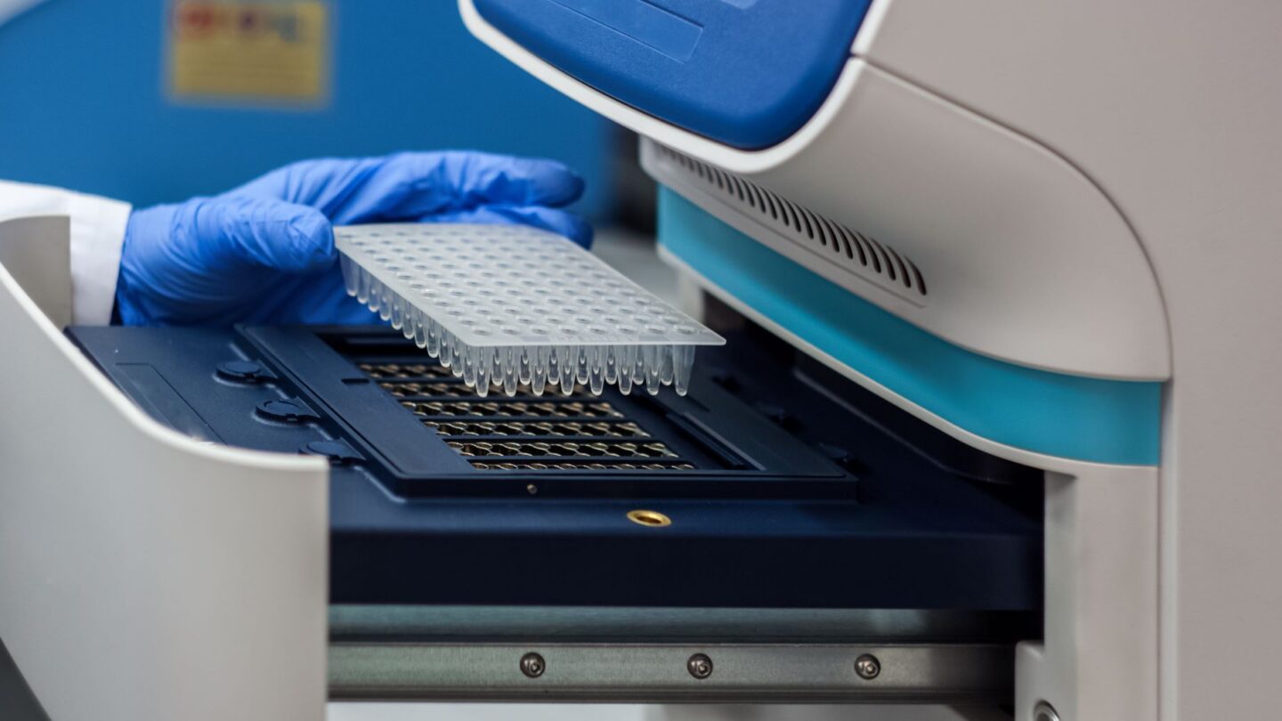 A photograph of DNA samples being placed in a PCR Machine by a gloved hand.