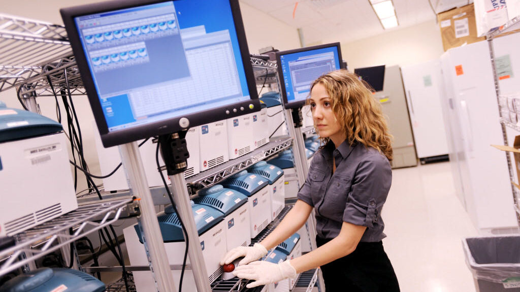 A photograph of Geneticist Pardis Sabeti working in a laboratory. She is standing beside a wheeled set of shelves holding multiple square containers. She is also looking towards a computer monitor sitting close to the shelves and her gloved hands are poised on a device with a big red button.