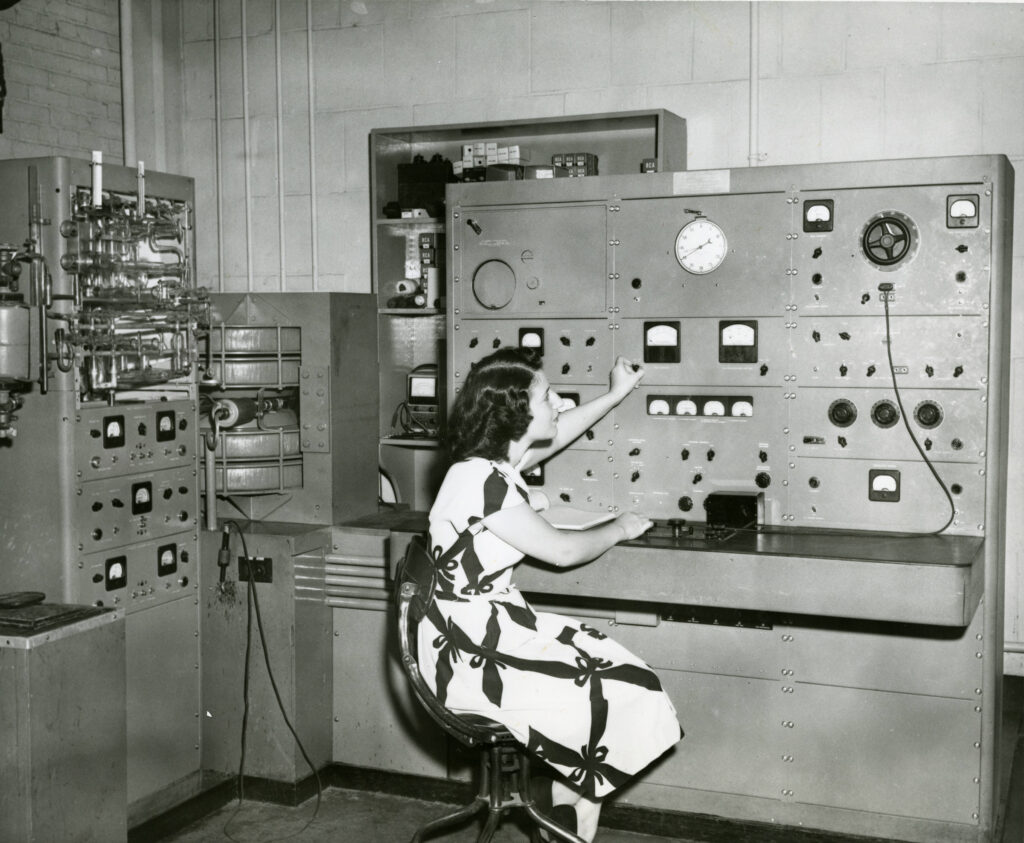 A woman in a patterned dress sits at a large, vintage control panel with dials, switches, and gauges in a laboratory or industrial setting.