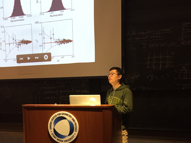 Man presenting slideshow with four graphs on it as he stands behind a lectern that says Simons Center for Geometry and Physics Stony Brook University