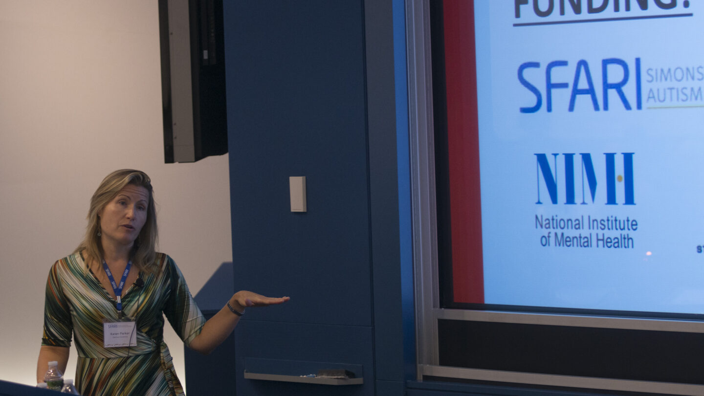 Woman behind a lectern, presenting a slideshow that has SFARI and NIMH logos