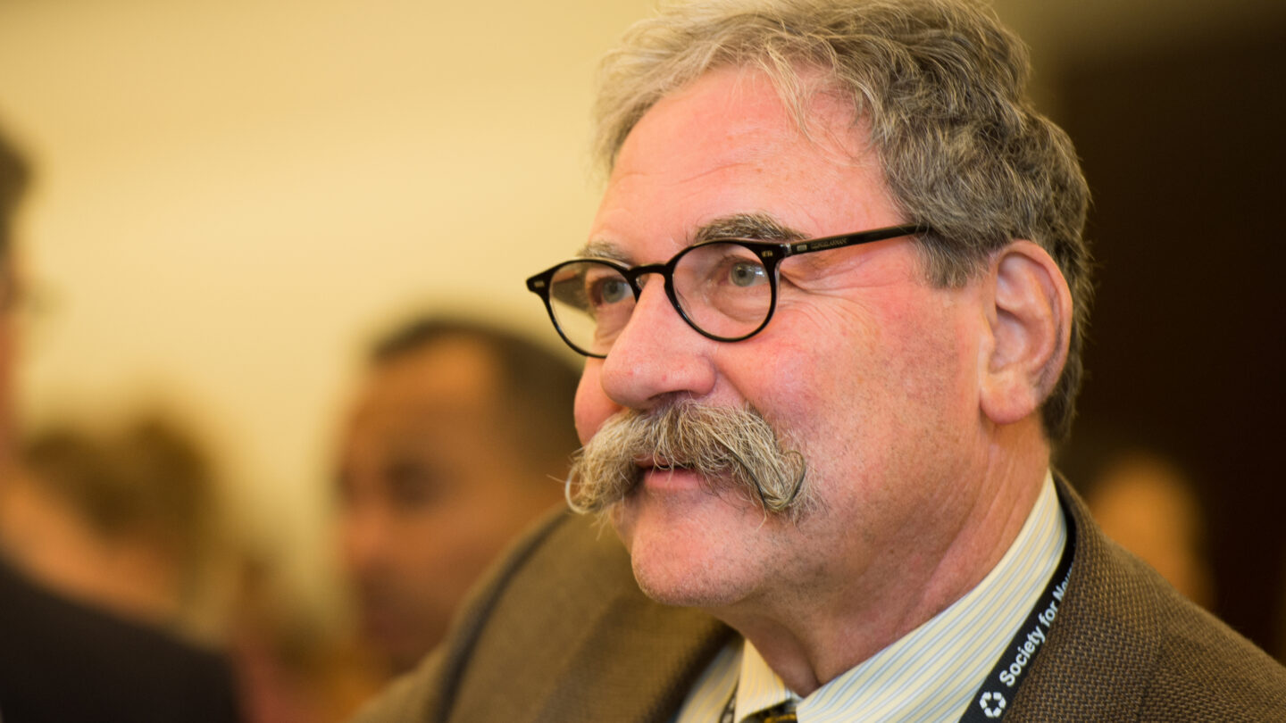An older man with gray hair, glasses, and a large mustache wears a brown suit and a lanyard. He is indoors and speaking, with blurred people and a yellow background behind him.