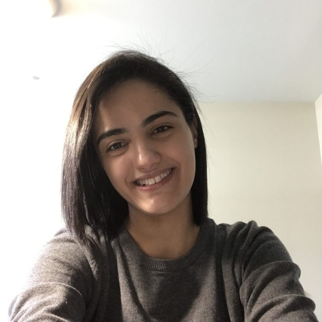 A person with straight, dark hair and a gray sweater smiles at the camera while sitting indoors in a well-lit room.