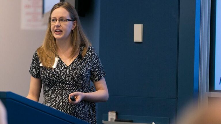 Lisa Giacomo of Stanford University stands behind a blue lectern as he presents a slideshow