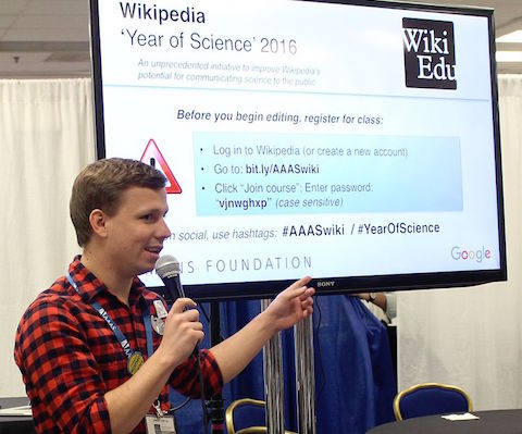 A person with a microphone points to a large screen displaying Wikipedia Year of Science 2016 instructions at an event, with registration details and hashtags visible on the screen.
