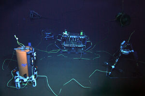 Three underwater robotic devices are connected by glowing cables in a dark, deep-sea environment, with various technical instruments visible around them.
