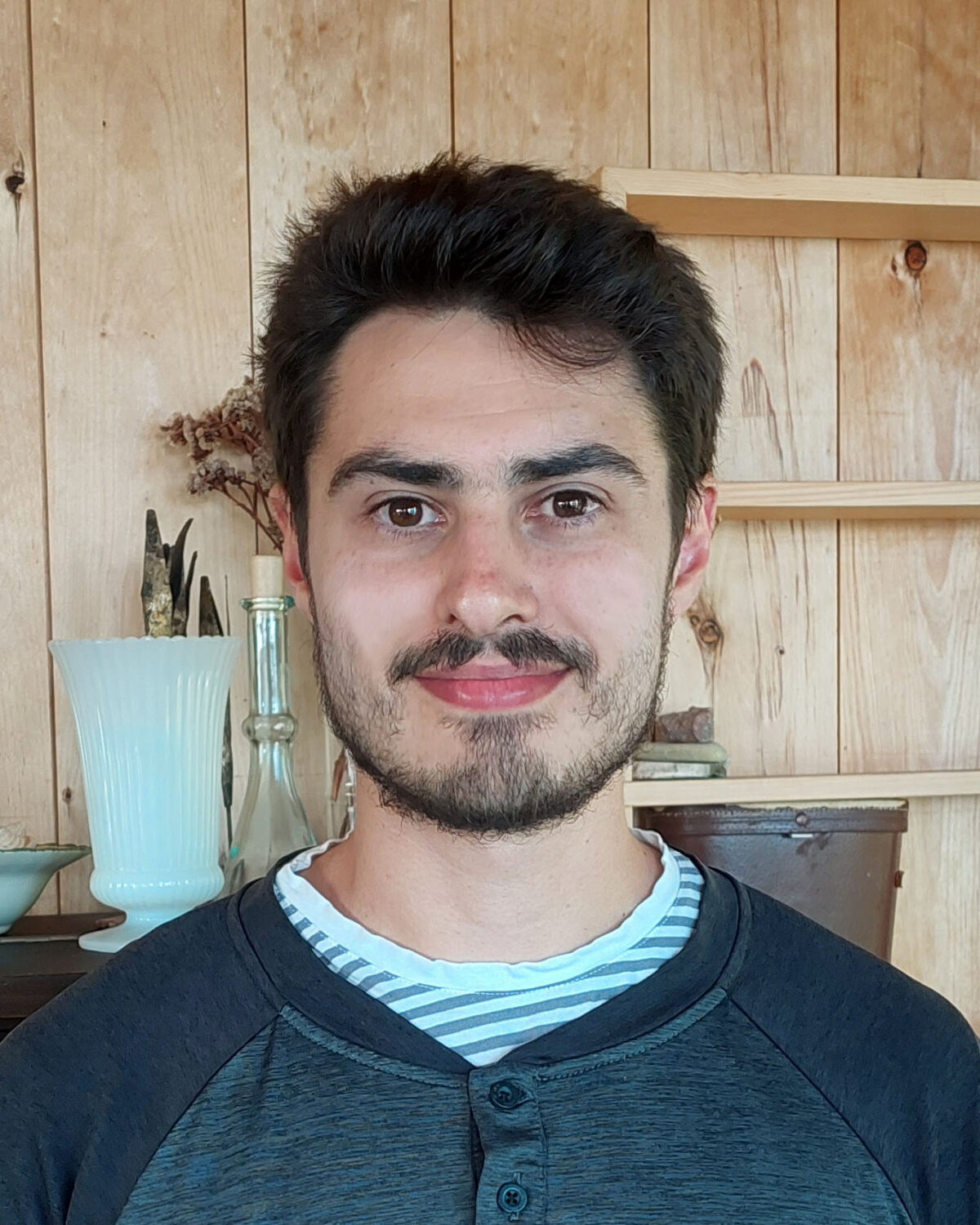 A young man with short dark hair and a trimmed beard smiles indoors. He is wearing a striped shirt under a dark buttoned shirt. The background has wooden paneling, shelves, and decorative items.