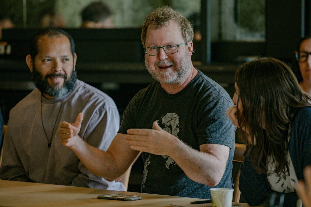 A man with glasses gestures while talking at a table with others. Two people sit beside him, listening attentively. A phone and a paper cup are on the table. The setting appears to be a meeting or discussion.