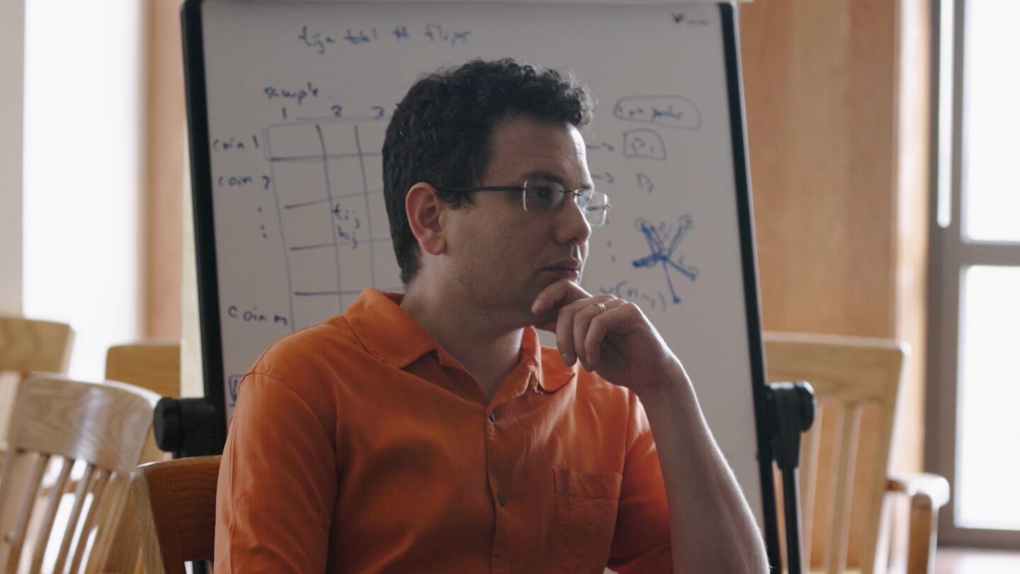 A person in an orange shirt sits thoughtfully with a hand on their chin in front of a whiteboard covered with diagrams and handwritten notes.