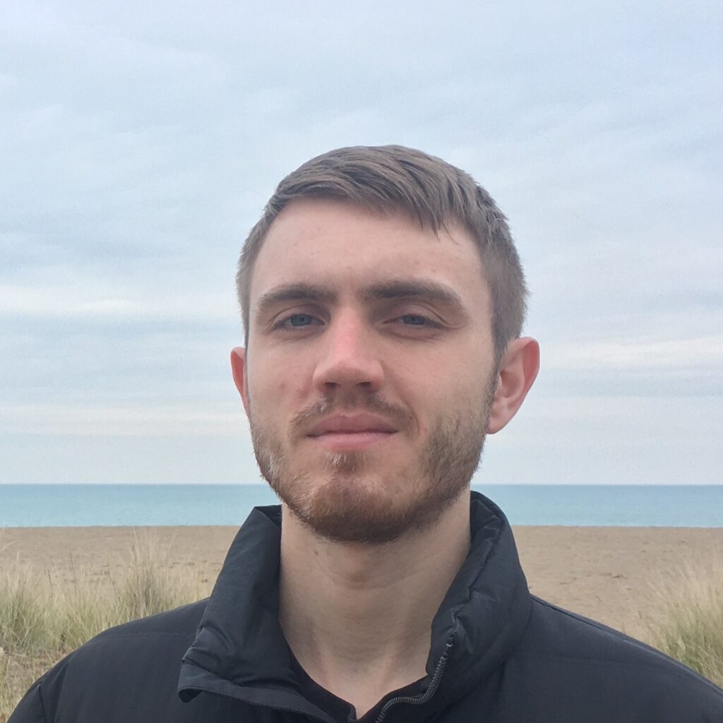 A young man with short brown hair and a beard stands outdoors on a cloudy day, with sand dunes, grass, and the sea in the background.