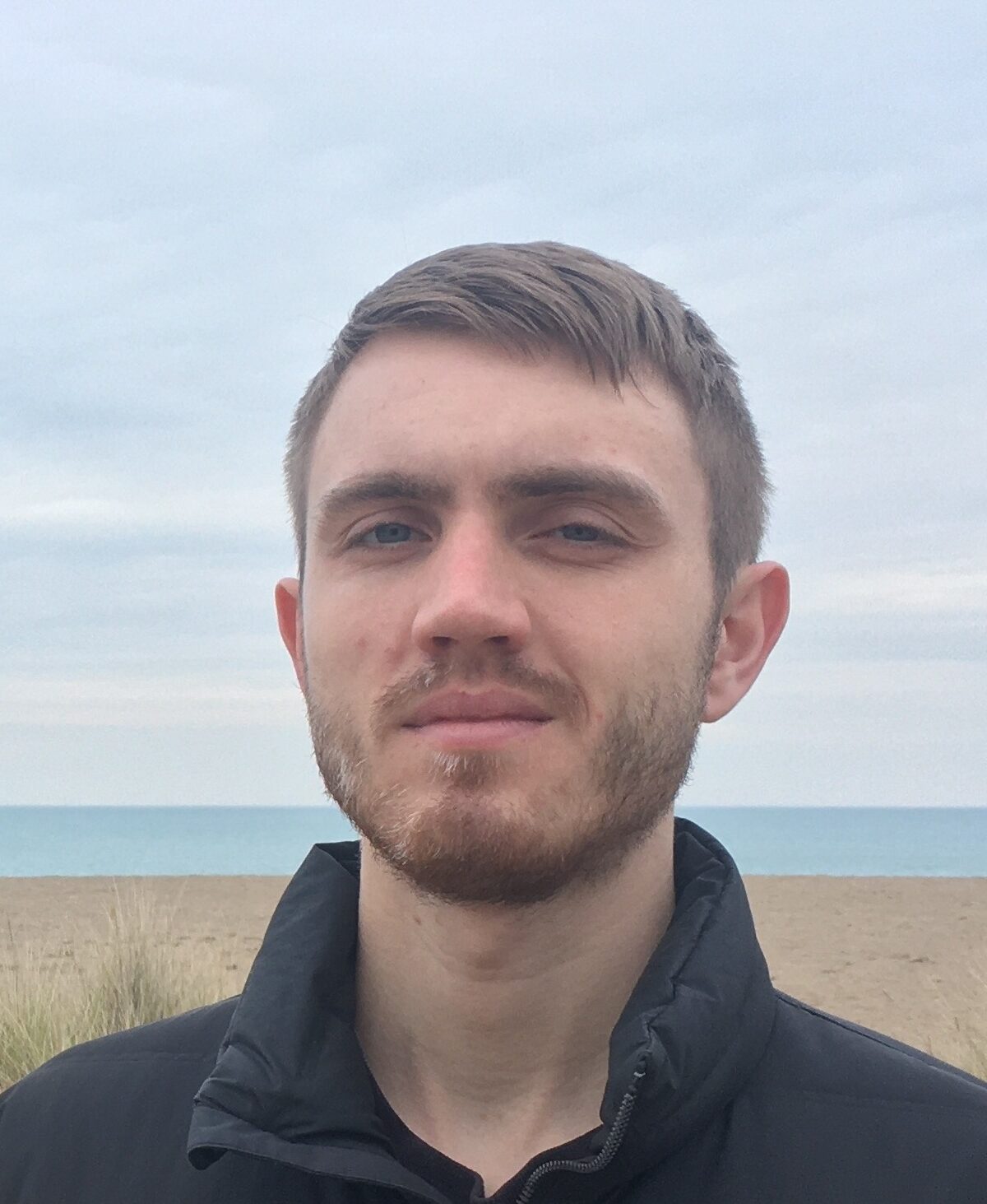 A young man with short brown hair and a beard stands outdoors on a cloudy day, with sand dunes, grass, and the sea in the background.