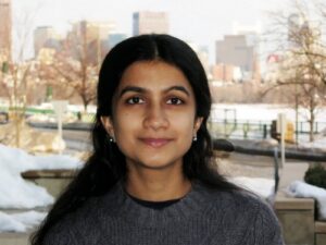 A woman with long dark hair wearing a gray sweater stands outside on a snowy day, with a blurred city skyline and bare trees in the background.