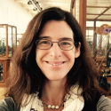 A woman with long brown hair and glasses smiles at the camera indoors, with wooden display cases and bright lighting in the background.