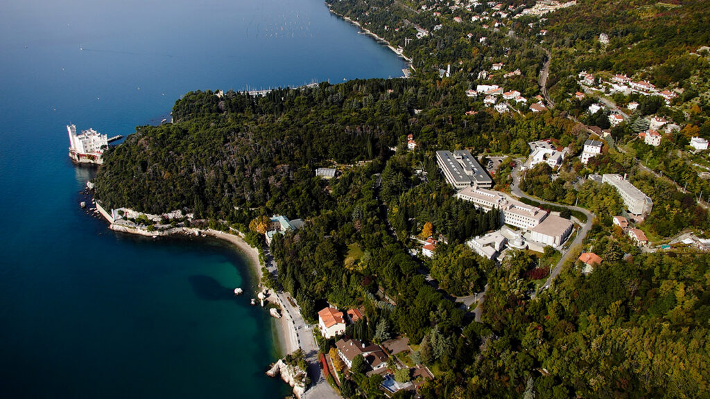 Aerial view of a lush, green coastline with scattered buildings, winding roads, and a docked ship. The land borders clear blue water, with the coastline curving gently and dense forest throughout.