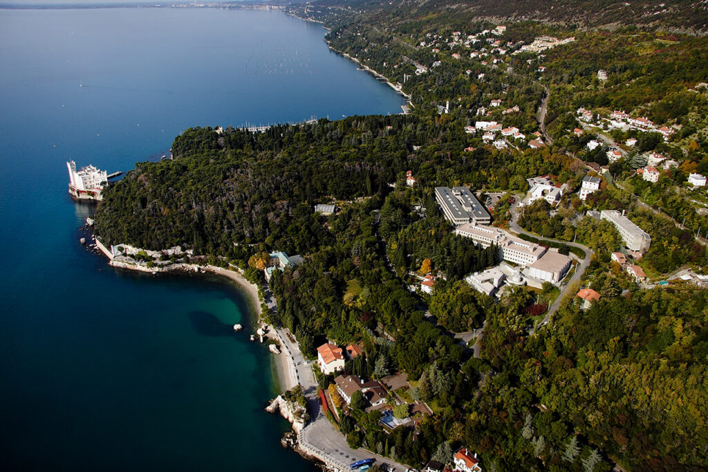 Aerial view of a lush, green coastline with scattered buildings, winding roads, and a docked ship. The land borders clear blue water, with the coastline curving gently and dense forest throughout.