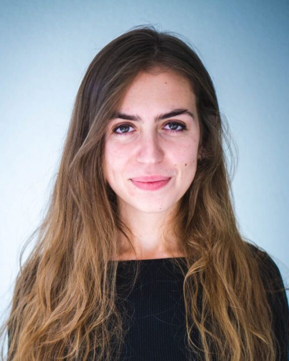 A young woman with long brown hair and a black top stands against a light blue background, looking at the camera with a neutral expression.