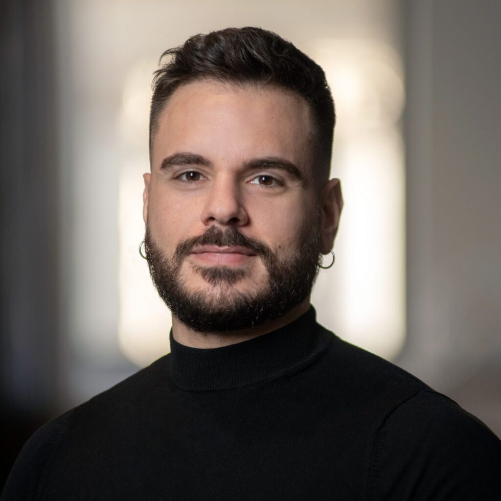 A man with short dark hair, a beard, and hoop earrings, wearing a black turtleneck, stands indoors with a neutral expression and a blurred background.