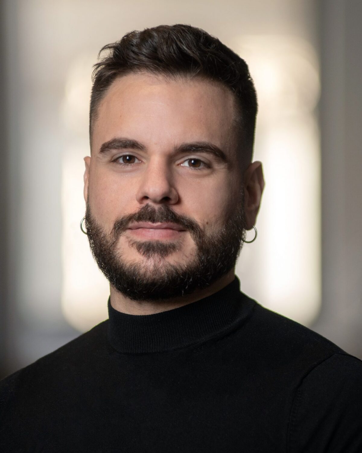 A man with short dark hair, a beard, and hoop earrings, wearing a black turtleneck, stands indoors with a neutral expression and a blurred background.